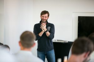 Dirk Primbs speaking to a room, hands together, the backs of listeners' heads in the foreground.