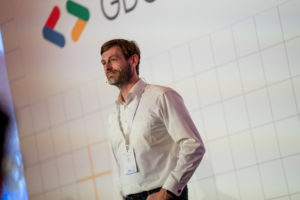 Dirk Primbs in a white shirt and lanyard, speaking in front of a wall carrying the Google logo.