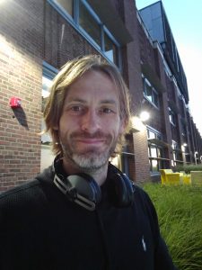 Portrait of Dirk Primbs outside a brick building, headphones around his neck, smiling at the camera.