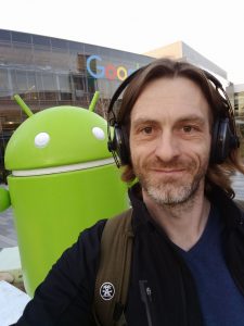 Dirk Primbs wearing over-ear headphones, standing in front of a large green Android statue.