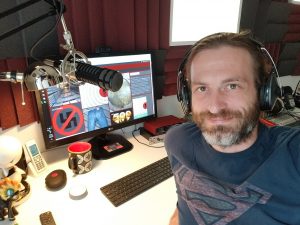 Dirk Primbs at a podcasting desk wearing headphones, with a studio microphone, monitor and keyboard.